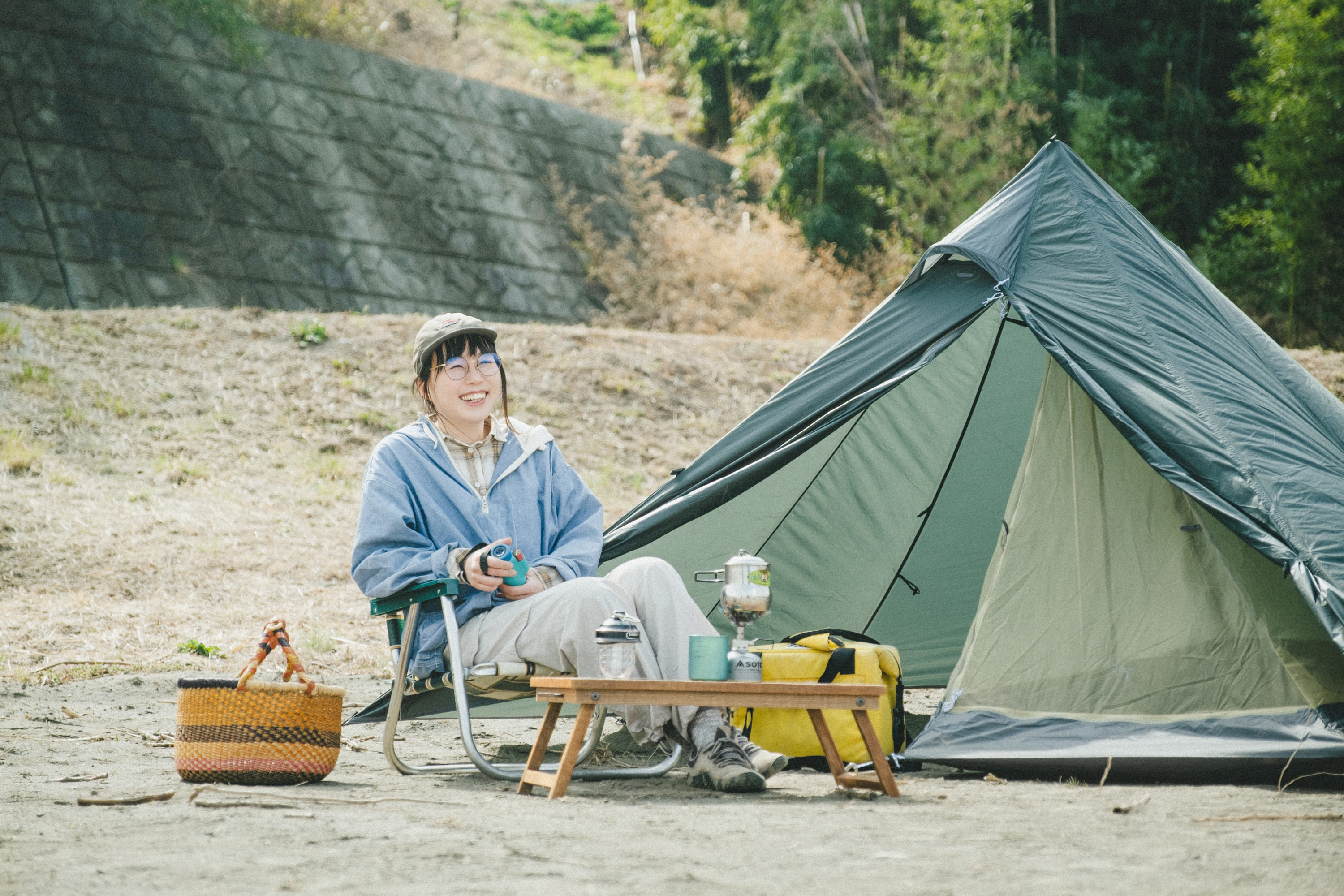 屋外のキャンプ場で、こいしさんがテントの前に置かれたアウトドアチェアに座って笑顔を見せている写真。