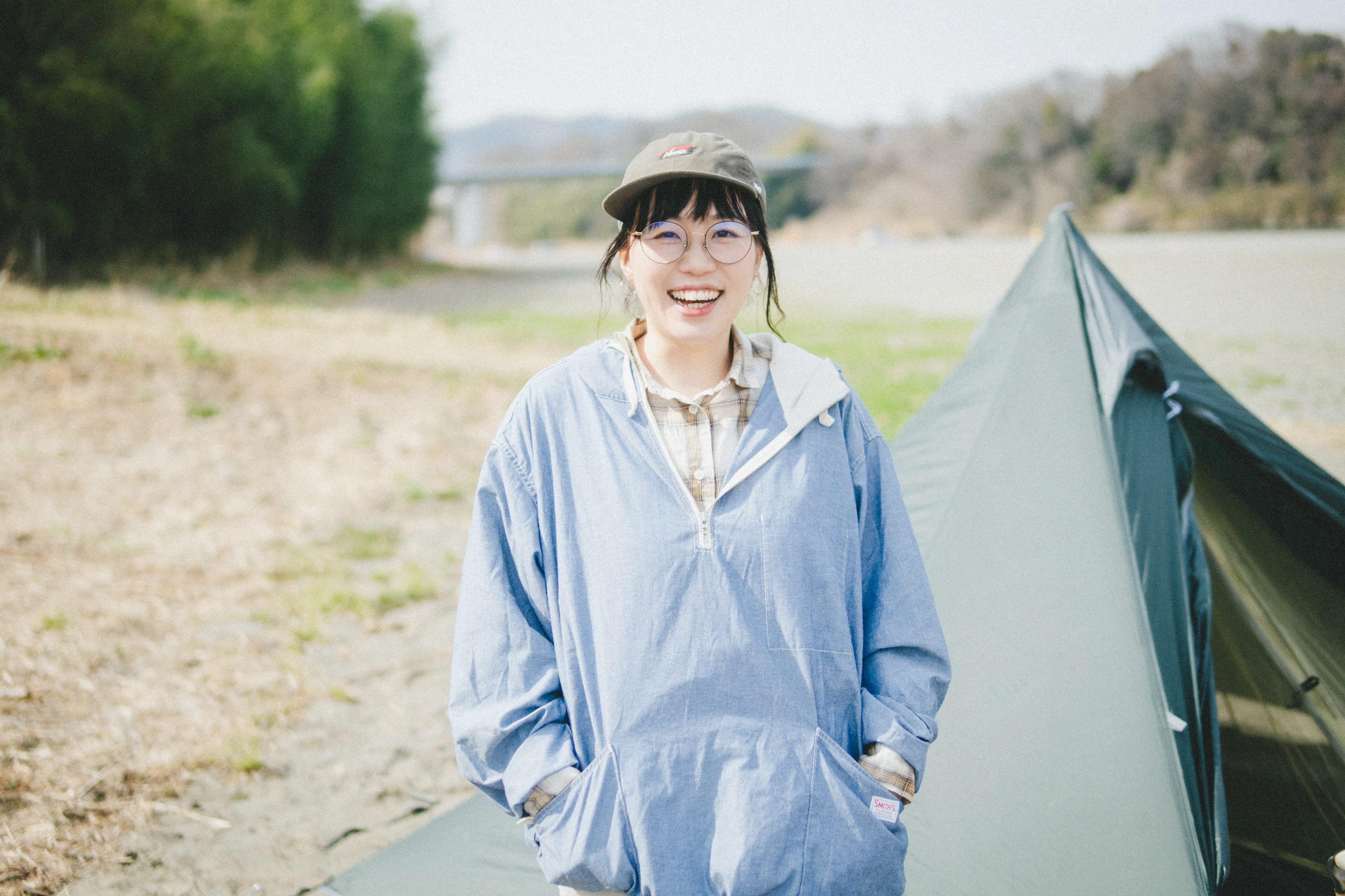 屋外のキャンプ場で、テントの横に立つこいしさんの上半身を写した写真。両手を上着のポケットに入れながら、正面を向いて大きく笑っている。隣には緑色のテントが設営され、背後には川や橋、木々が広がる自然の風景が見える。