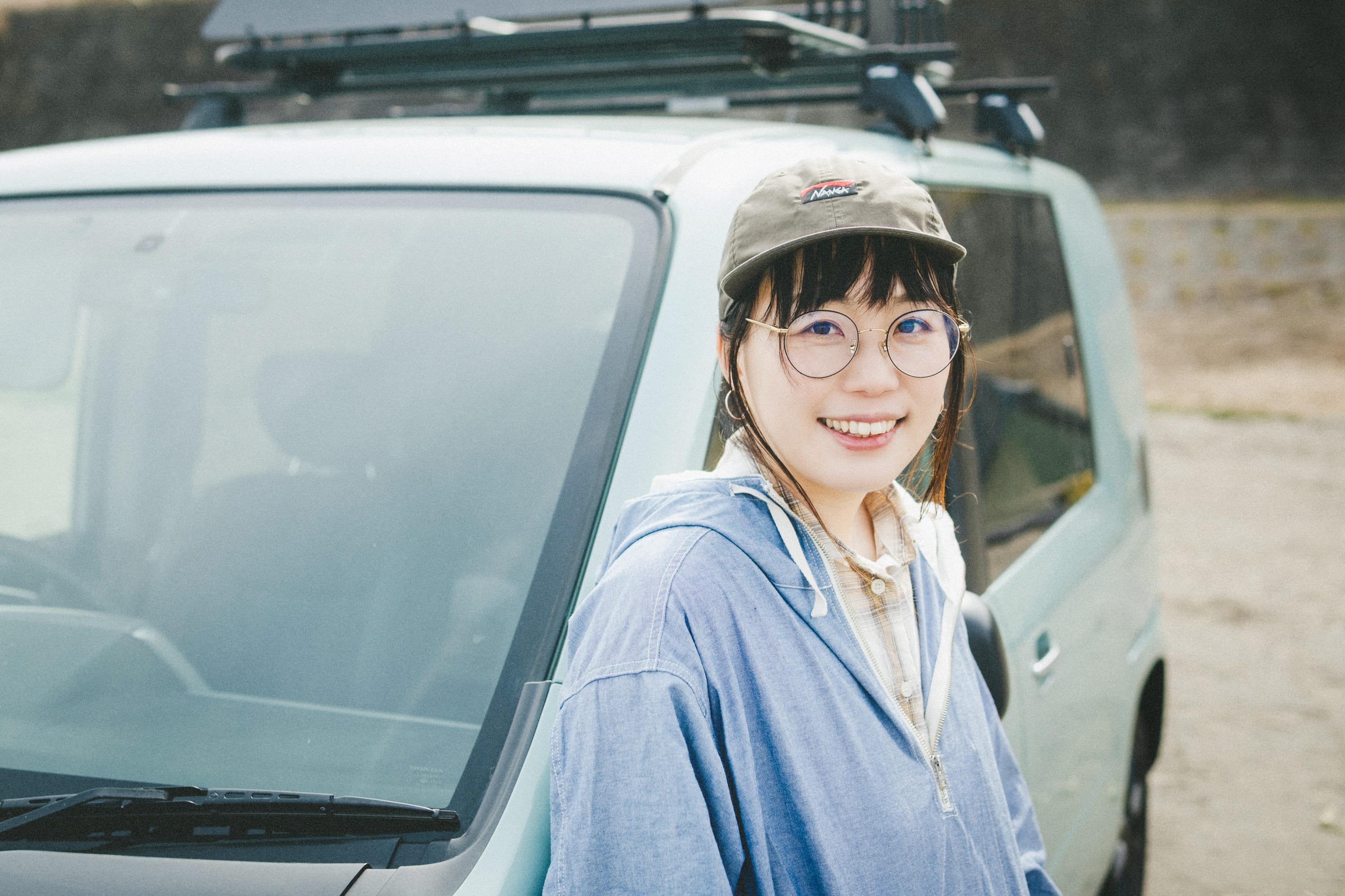 車の前に立つこいしさんの写真。背後には淡いグリーンの車があり、屋根にはルーフキャリアが取り付けられている。背景は屋外の空き地のような場所で、落ち着いたアウトドアの雰囲気が感じられる情景。