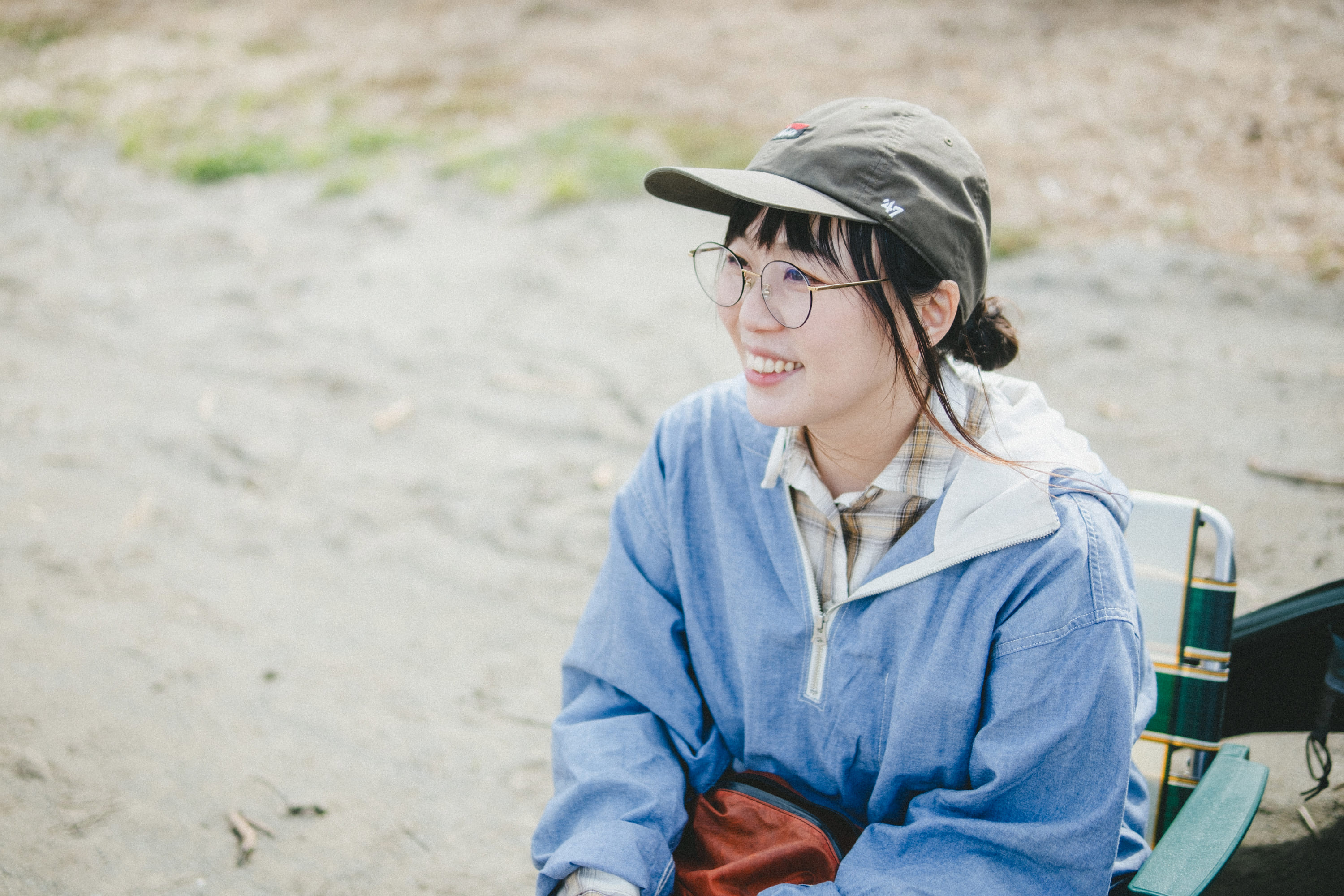 屋外のキャンプ場で、アウトドアチェアに座ったこいしさんの写真。背景には砂地や草地が広がり、自然の中でくつろいでいる様子が伝わる情景である。
