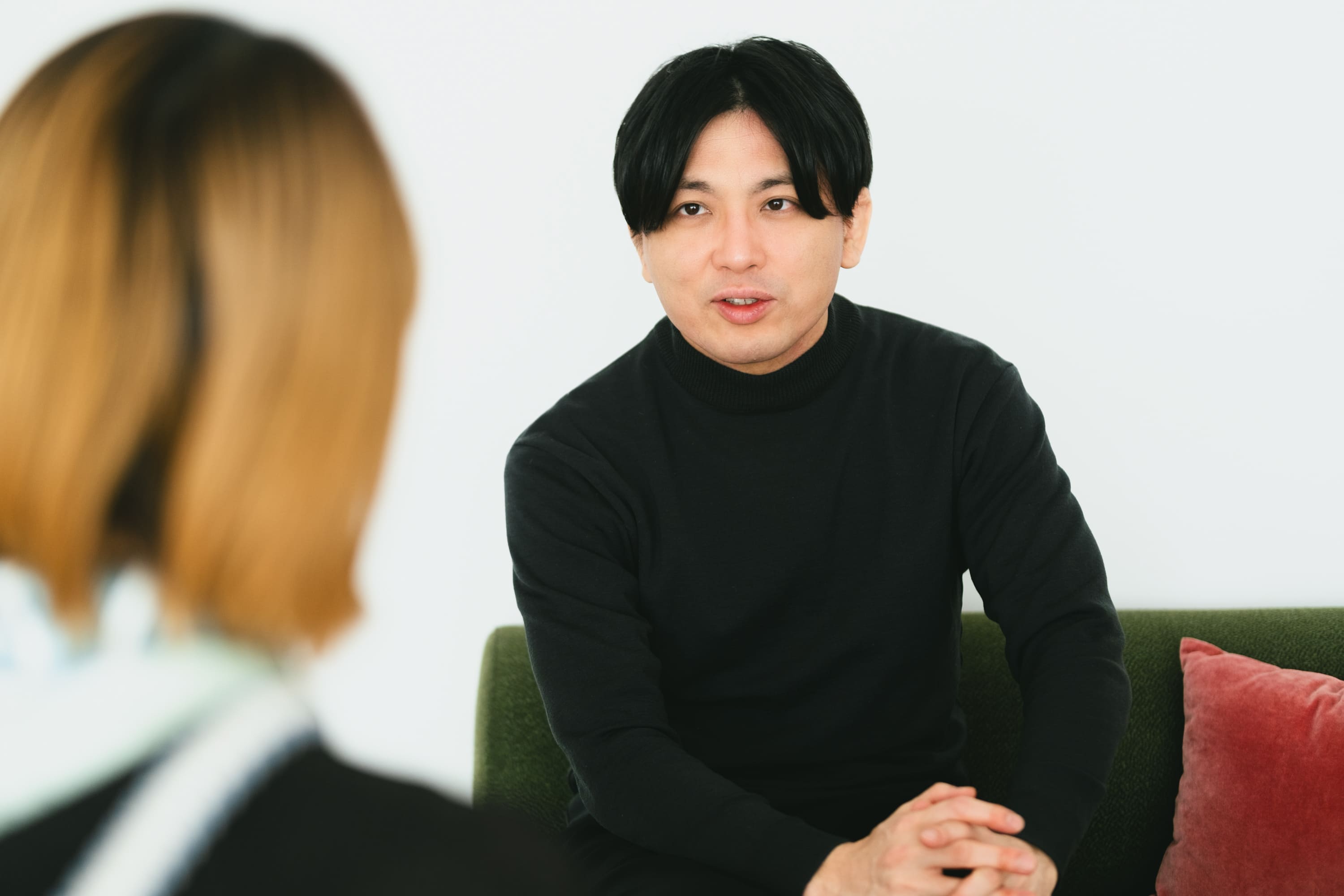 益田さんのバストショット。緑色のソファに腰掛け、体をやや前に傾けながら向かい側の人物に視線を向けている。両手を軽く組み、真剣な表情で話を聞いている。