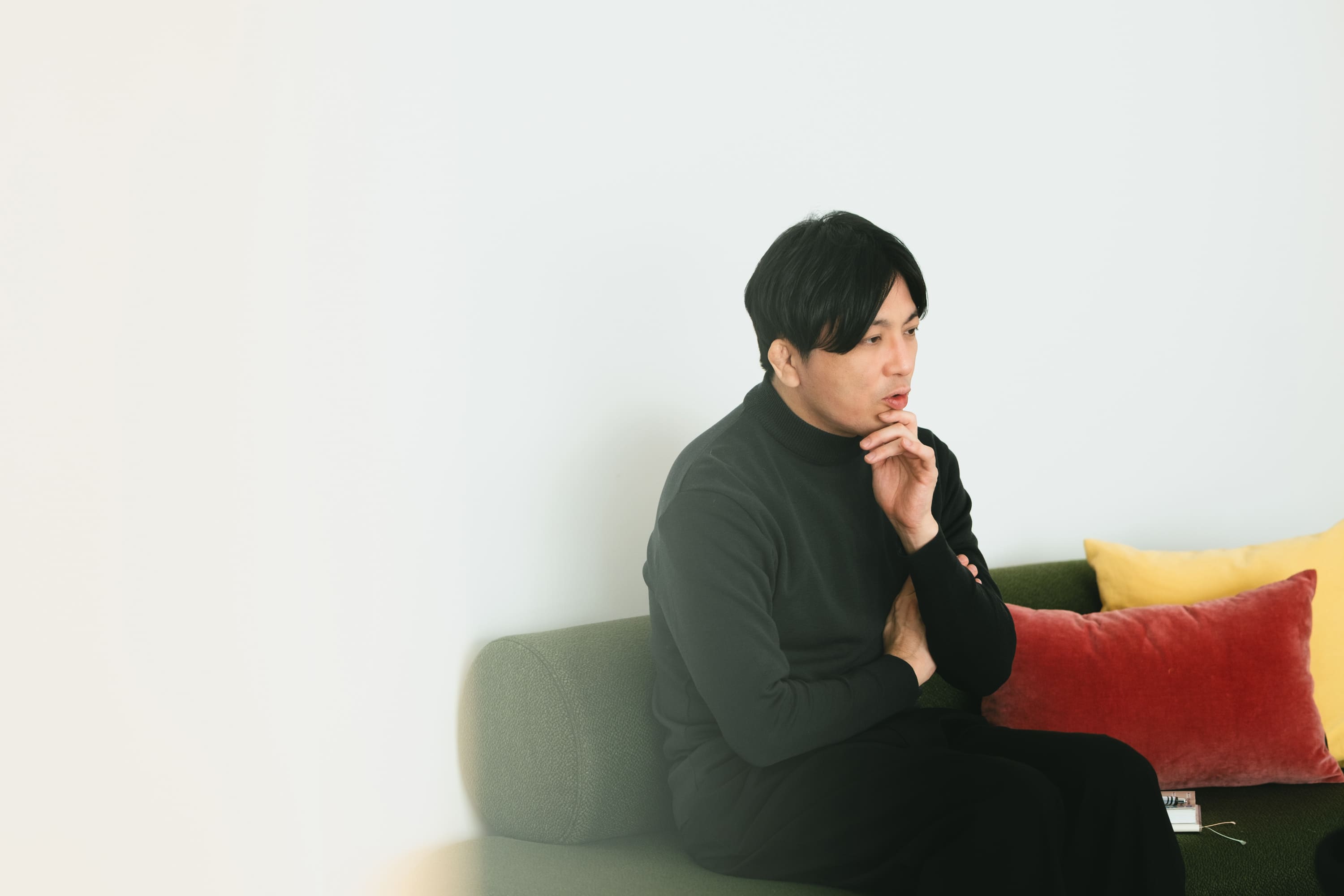 益田さんのバストショット。緑色のソファに腰掛け、体をやや前に傾けている。右手の指をあごに当て、左腕を胸の前で抱えるようにしており、視線を下方に向けて思案する表情をしている。