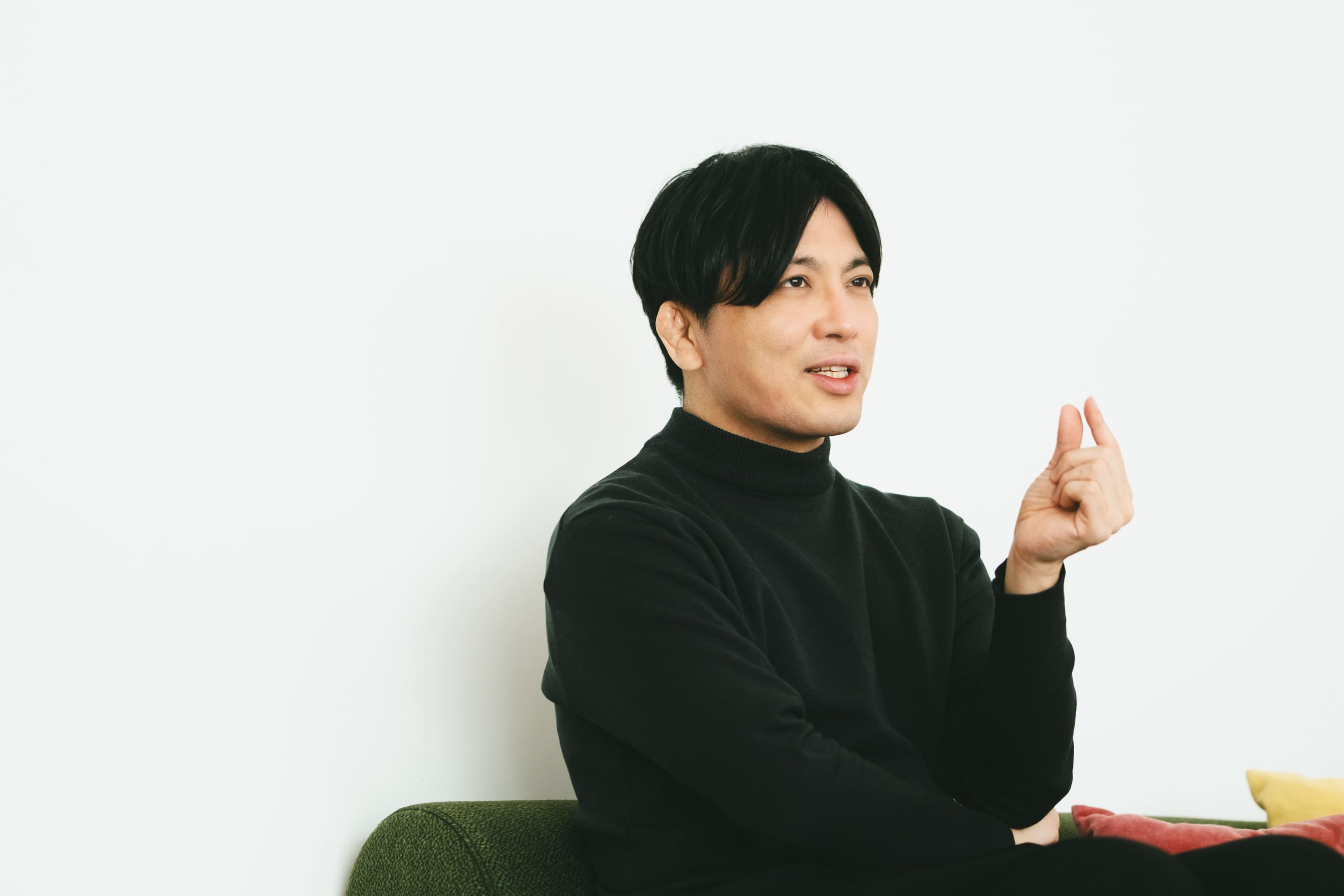 益田さんのバストショット。緑色のソファに腰掛け、体を横向きにしながら話している様子である。右手の親指と人差し指を軽く近づけて上げ、説明するような仕草をしている。