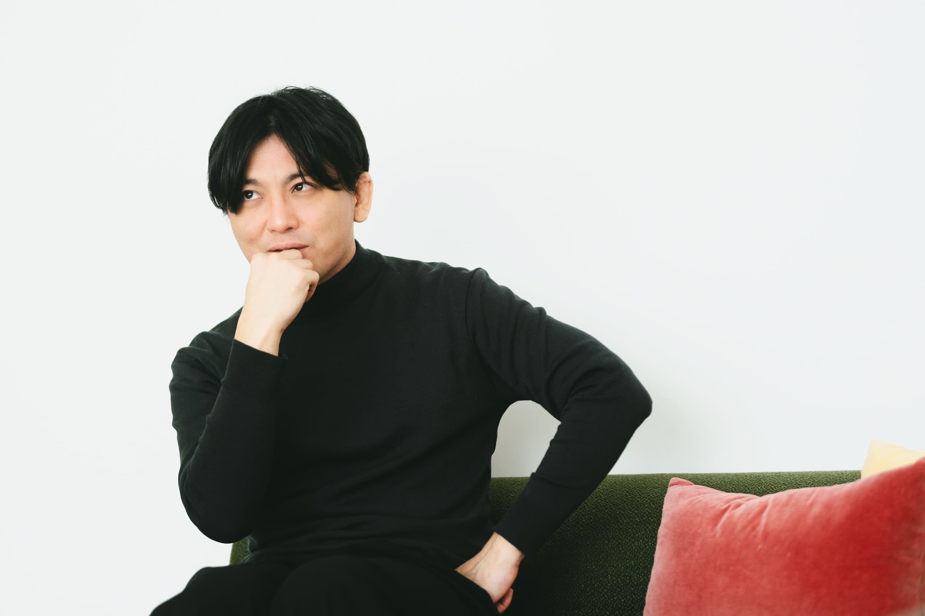 益田さんのバストショット。緑色のソファに腰掛け、右手を口元に当てて軽く握り、左手を腰に添えている。視線をやや上に向け、考え込むような表情をしている。