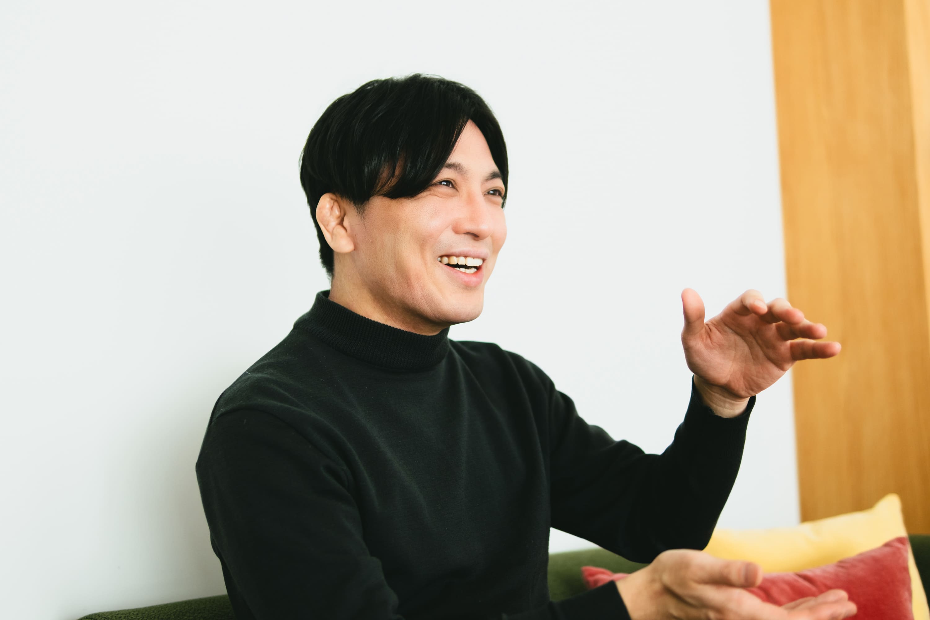 益田さんのバストショット。右手を上げて指を軽く曲げ、身振りを交えて話しており、左手も前に出している。背景には白い壁と木目の柱、黄色や赤のクッションが見える。