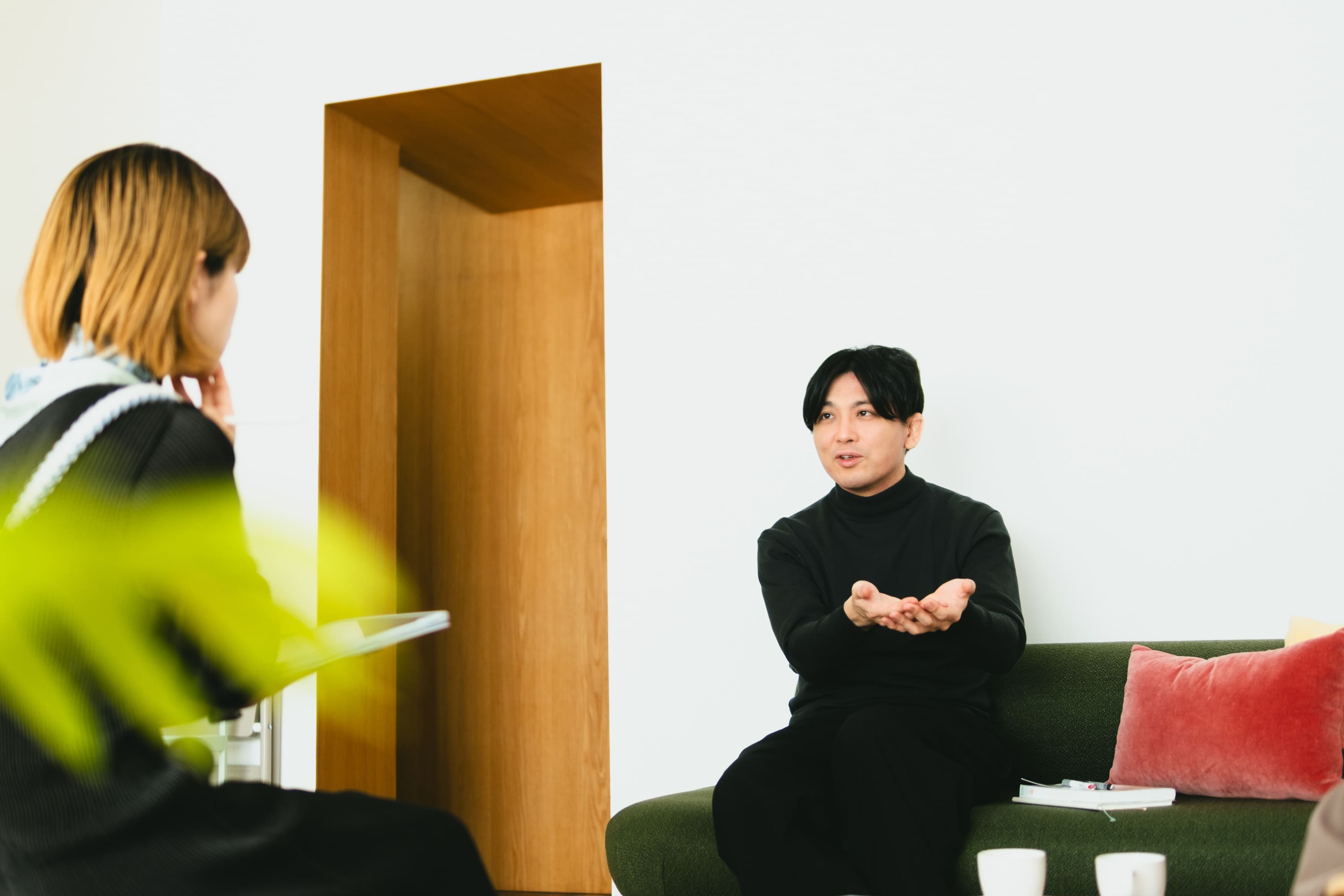 :益田さんのバストショット。緑色のソファに腰掛け、向かい側の人物に向かって両手のひらを上に向けて差し出しながら話している。体をやや前に傾け、説明するような穏やかな表情をしている。手前には後ろ姿の人物が写っており、背景には白い壁と木目の開口部、赤いクッションが見える。