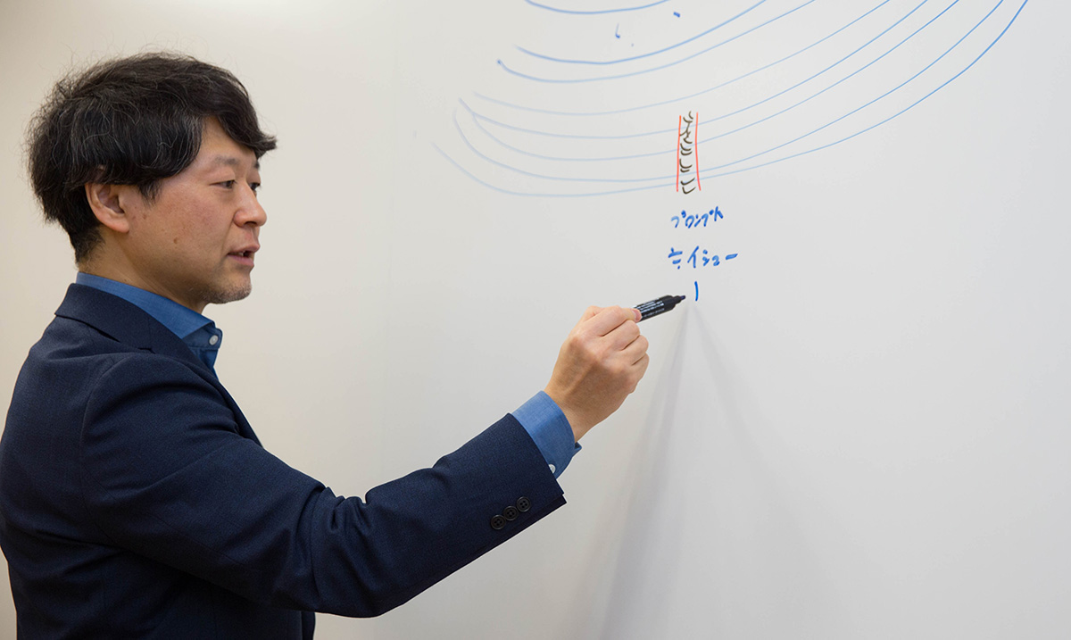 浦崎がホワイトボードに向かい、黒いマーカーで文字を書いている様子。青と赤の線で構成された図の下に「プロジェクト」「イシュー」と書き込みながら、考えを整理している。