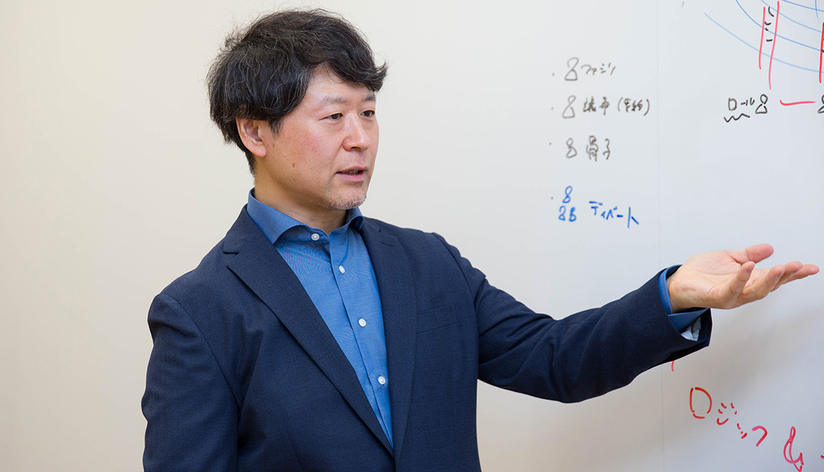 浦崎がホワイトボードの前で右手を広げ、説明している様子。ボードには青と赤のマーカーで図や文字が書かれている。