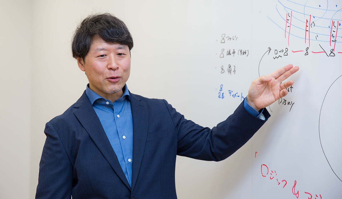 浦崎がホワイトボードの前で右手を上げ、指し示しながら話している様子。