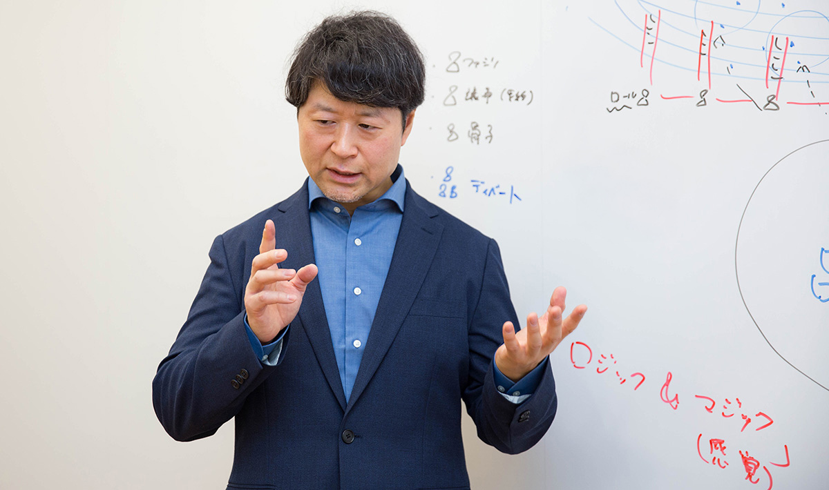 浦崎がホワイトボードの前で両手を使って話をしている様子。視線は下向きで考えながら説明しているように見える。ボードには赤と青のマーカーで図や文字が描かれている。