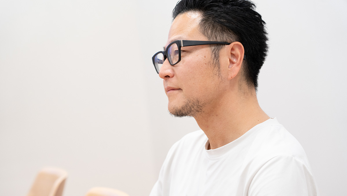 メガネをかけた男性が横を向いて座っている様子です。白い背景で真剣な表情です。