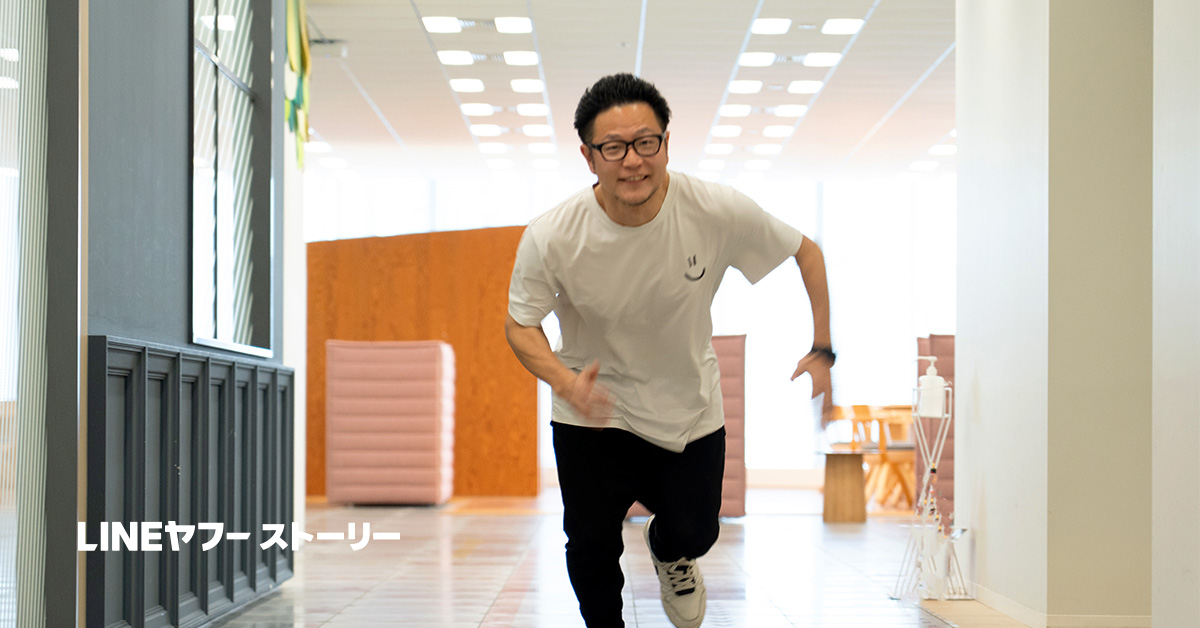 白いTシャツを着た男性が、オフィスの廊下を走っている様子です。