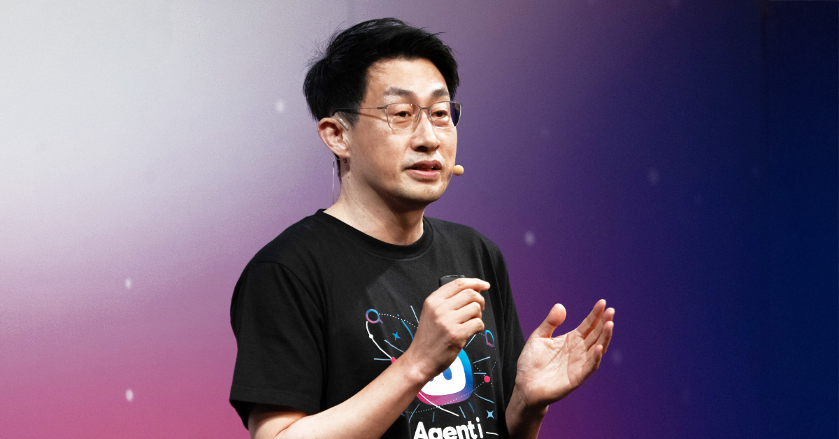 慎がステージ上でプレゼンテーションをしている写真。黒い「Agent i」とロボット風アイコンがデザインされたTシャツを着用し、右手に小型のリモコンを持ち、左手を軽く開きながら説明している。