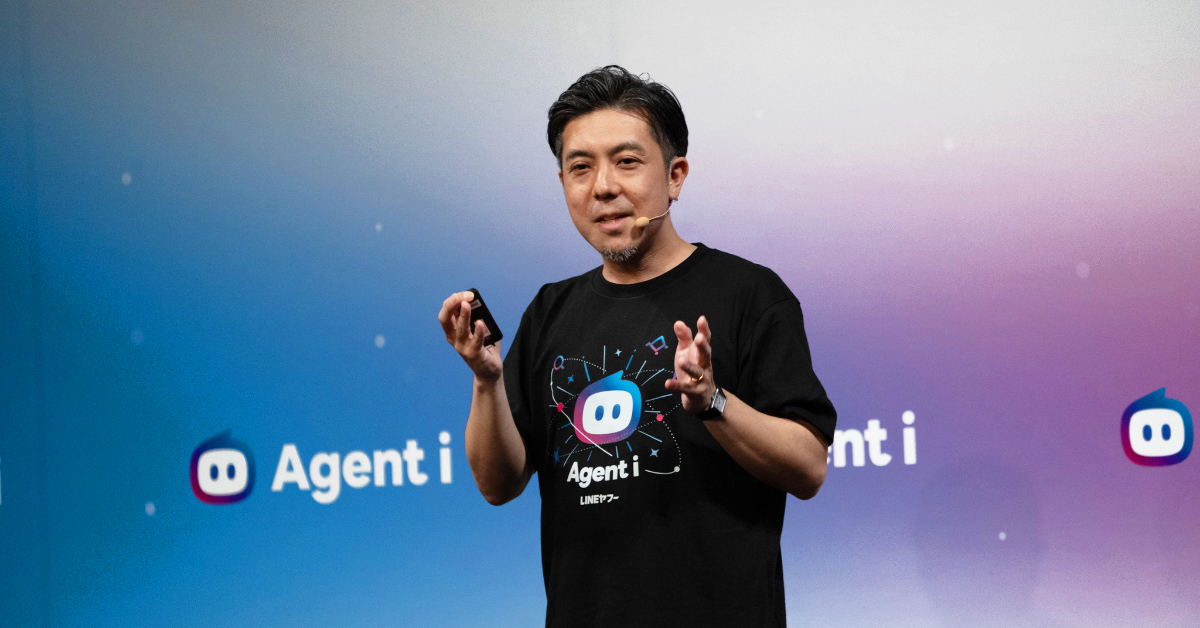 葭沢がステージ上でプレゼンテーションをしている写真。黒い「Agent i」とロボット風アイコンが描かれたTシャツを着用し、右手を前に出して指を広げながら説明する仕草をしている。