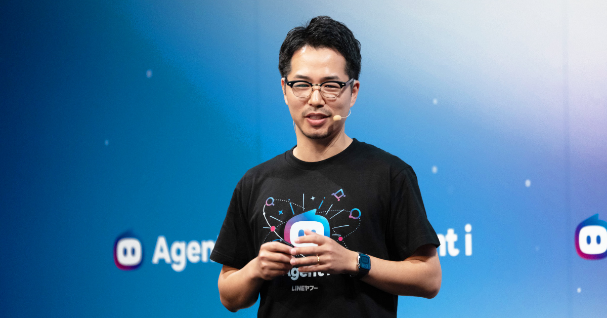 二木がステージ上でプレゼンテーションをしている写真。黒い「Agent i」とロボット風アイコンがデザインされたTシャツを着用し、両手で小型のリモコンを持ちながら話している。