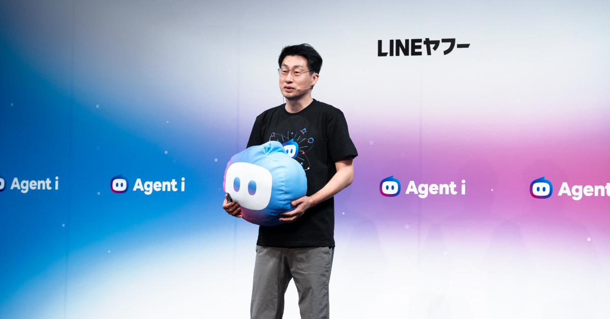 慎がステージ上でクッション状のキャラクターを両手で抱えながら話している写真。