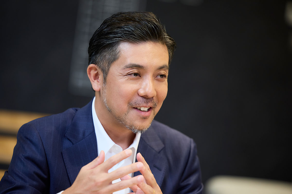 オフィスで、葭沢が顔の前で両手を組みながら話している写真。ネイビーのスーツを着て、柔らかな表情を浮かべている。