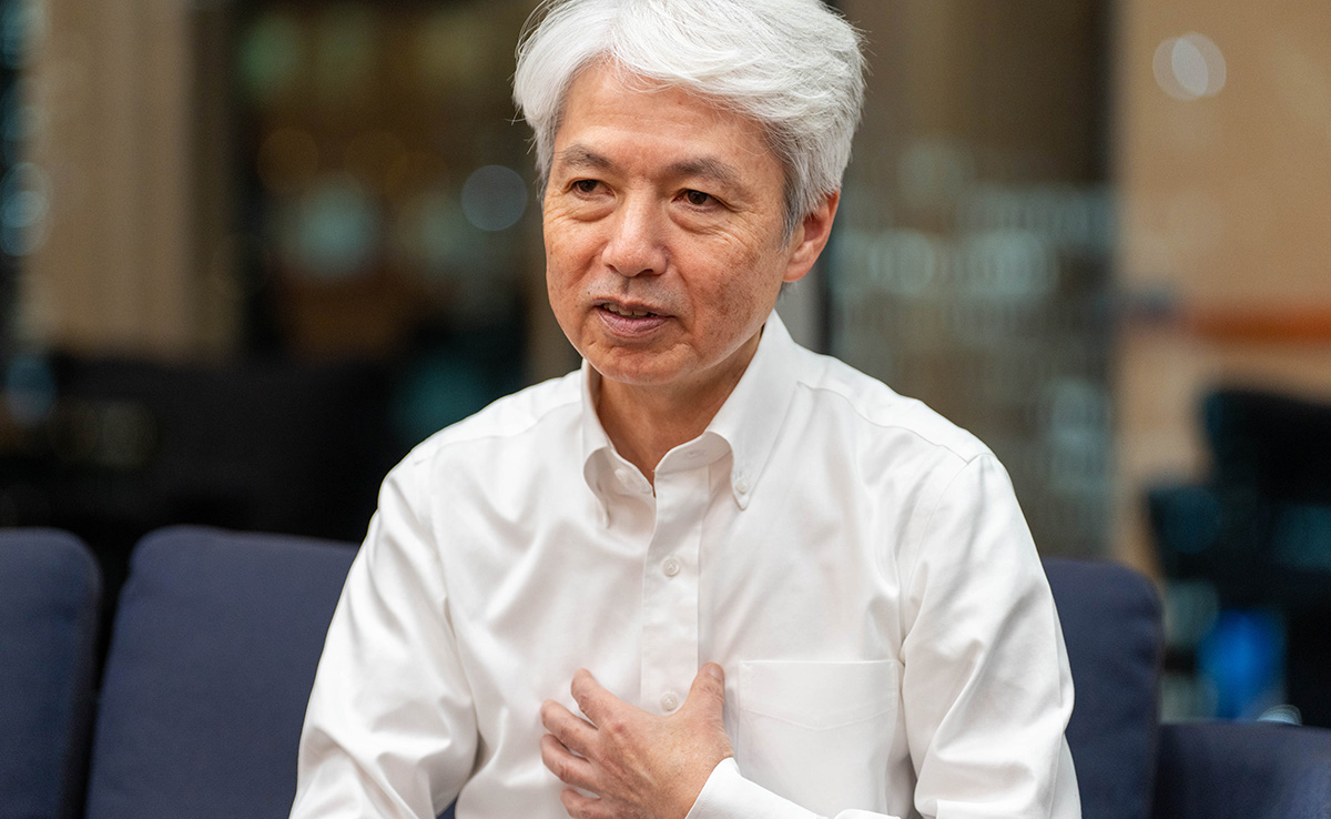 岩崎のバストショット。白髪のショートヘアで、白いワイシャツを着ている。ソファに座り、左手を胸に当てながら真剣な表情で前を見て話している様子。