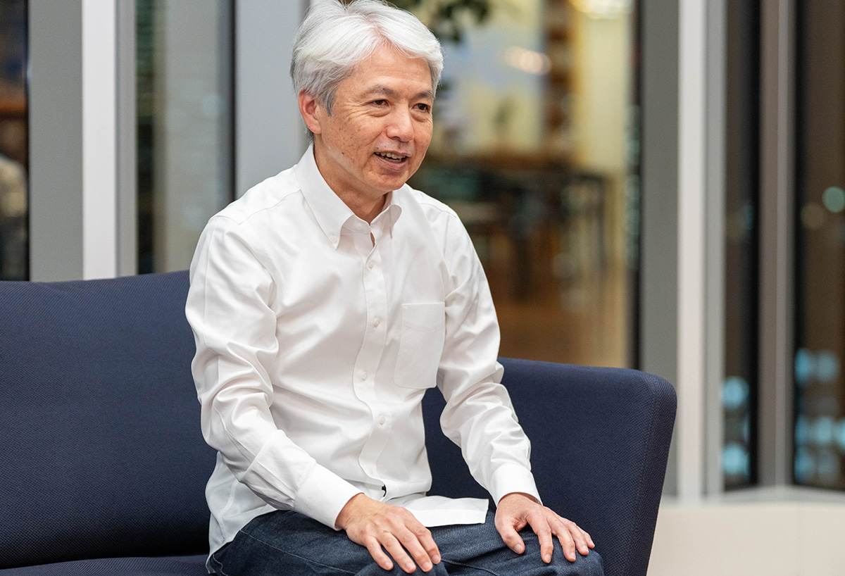 岩崎の上半身から膝上までを写したショット。白髪のショートヘアで、白いワイシャツと濃い色のズボンを着て、ネイビーのソファに座っている。軽く前かがみになりながら両手を膝の上に置き、笑顔で話している様子。