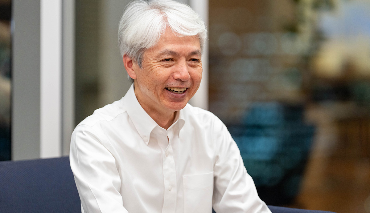 岩崎のバストショット。白髪のショートヘアで、白いワイシャツを着ている。ネイビーのソファに座り、笑顔でやや前傾姿勢になっている。