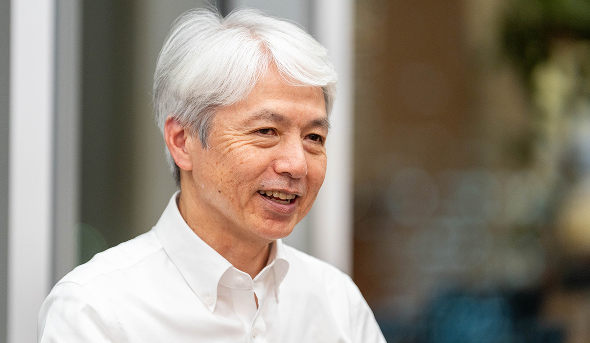 岩崎のバストショット。白髪のショートヘアで、白いワイシャツを着ている。少し体を右に向けながら笑顔で話している様子。