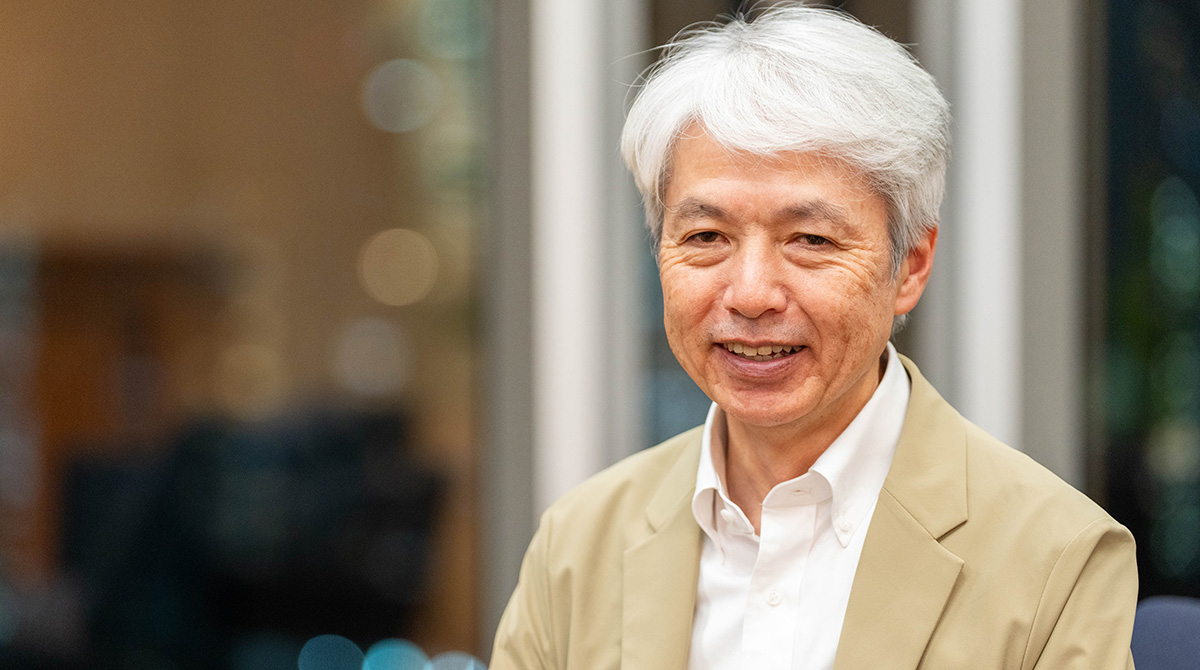 岩崎のバストショット。白髪のショートヘアで、白いワイシャツとベージュのジャケットを着ている。穏やかに微笑みながらカメラのほうを向いている。