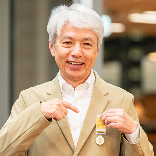 岩崎の写真。白髪のショートヘアで、白いワイシャツとベージュのジャケットを着ている。笑顔を見せながら、胸元につけた黄色のリボンとメダルを指差している。