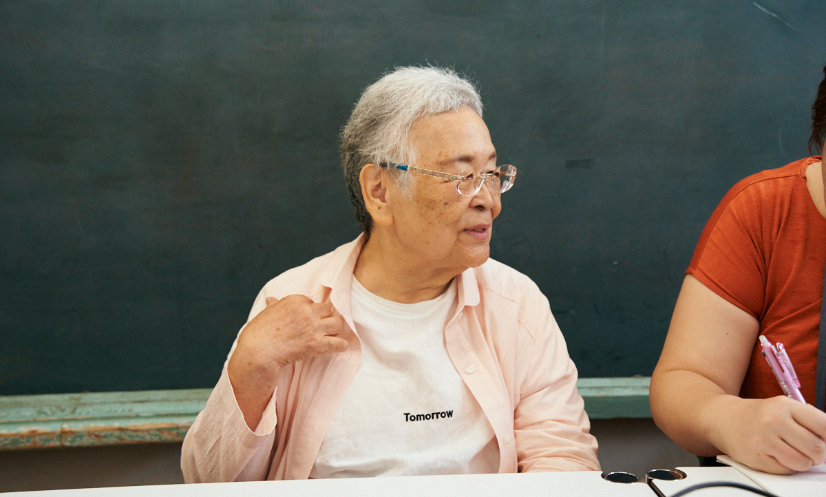シニアの女性が机に座り、隣の人に向かって話しかけている。白髪で眼鏡をかけ、白いTシャツの上に淡いピンクのシャツを羽織っている。左手で胸元に軽く触れながら、横を向いて穏やかに話している様子だ。右側にはオレンジ色の服を着た人物の腕が写っており、ペンを持って何かを書いている。背景には黒板がある。