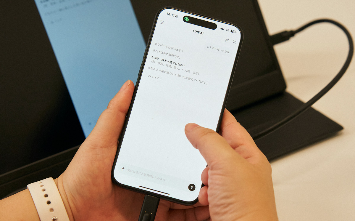 手に持ったスマートフォンの画面に「LINE AI」と表示され、会話のような文章が表示されている。画面には「ありがとうございます！」「それでは次の質問です」「その時、誰と一緒でしたか？」などの文字が読み取れる。スマートフォンはケーブルで黒いタブレット端末のような機器に接続されており、背景にその画面がぼんやり映っている。