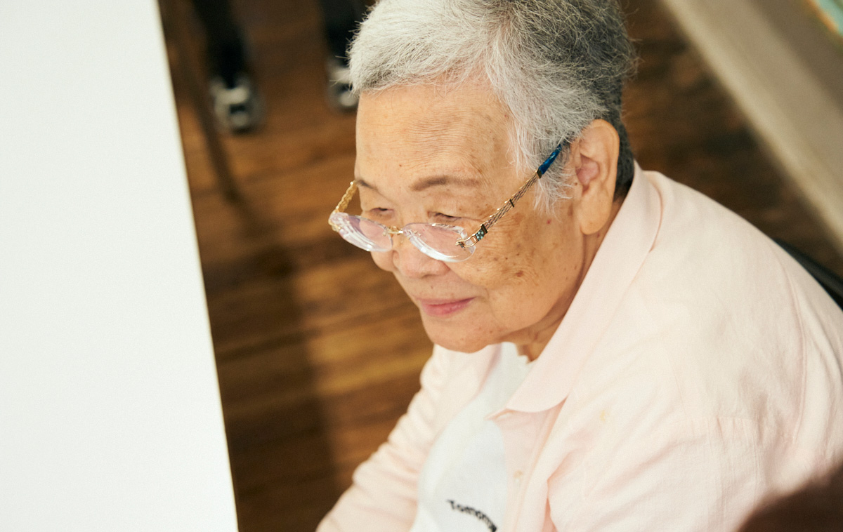 シニアの女性が机に向かって座り、やさしく微笑んでいる。白髪で眼鏡をかけ、白いTシャツの上に淡いピンクのシャツを羽織っている。木の床と白い机が写っており、明るい室内の光に包まれた穏やかな雰囲気の写真である。