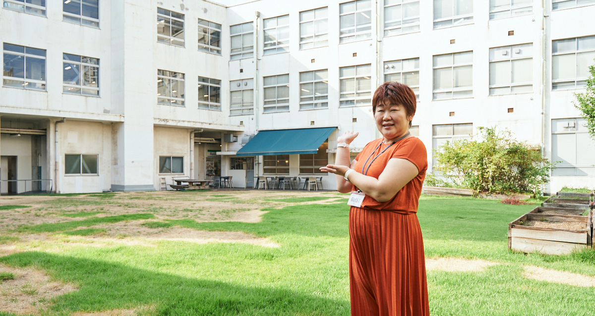 短髪でオレンジ色のワンピースを着た女性が、屋外の中庭のような場所で建物を指さして立っている。建物は白い外壁で複数の窓があり、学校や公共施設のような印象がある。中庭には緑の芝生と一部に枯れた草地が混ざり、右側には木製の花壇が並んでいる。女性は胸に名札を下げ、笑顔で建物を紹介しているように見える。