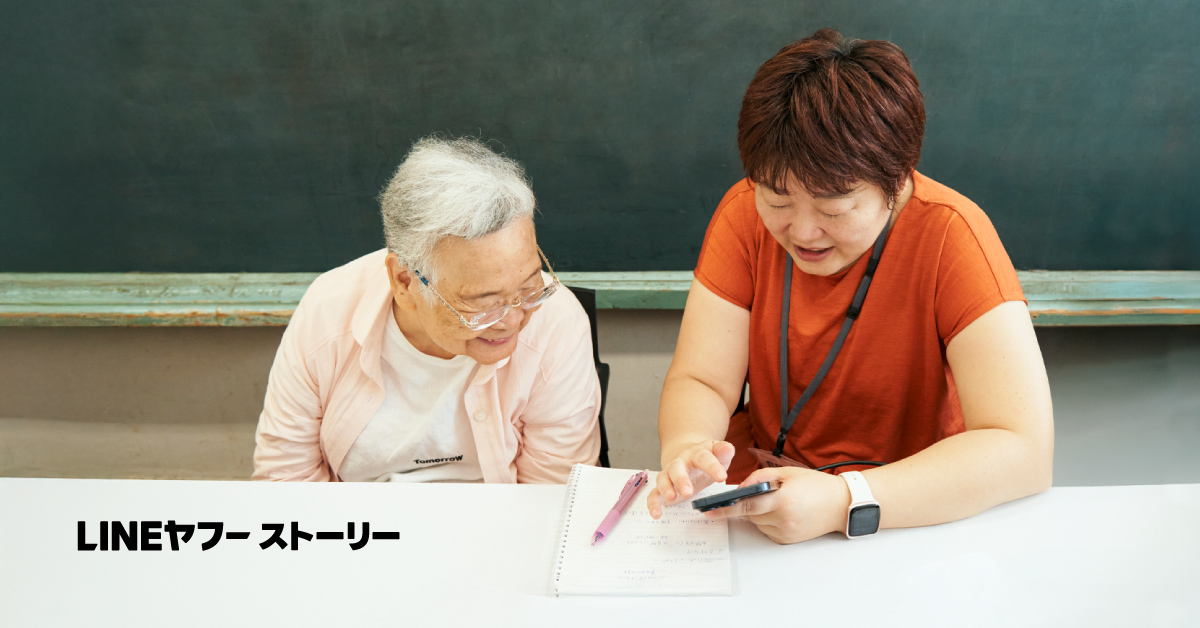 教室のような場所で、シニアの女性と短髪の女性が並んで座り、スマートフォンを見ながら会話している。シニアの女性は白髪で眼鏡をかけ、白いTシャツに淡いピンクのシャツを羽織っている。隣の女性はオレンジ色のTシャツを着ており、手にスマートフォンを持って操作している。2人とも笑顔で楽しそうな様子で、背景には黒板がある。左下に「LINEヤフー ストーリー」と書かれている。