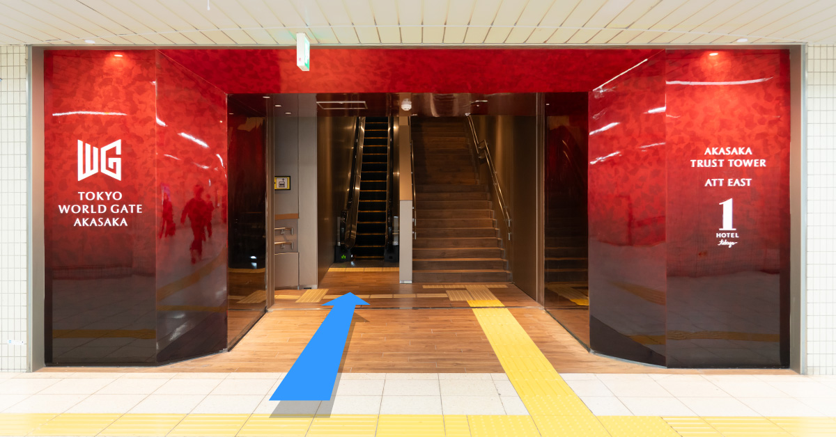 赤い壁面の入口「TOKYO WORLD GATE AKASAKA」「AKASAKA TRUST TOWER ATT EAST」。中央にエスカレーターと階段があり、青い矢印で進行方向を示す