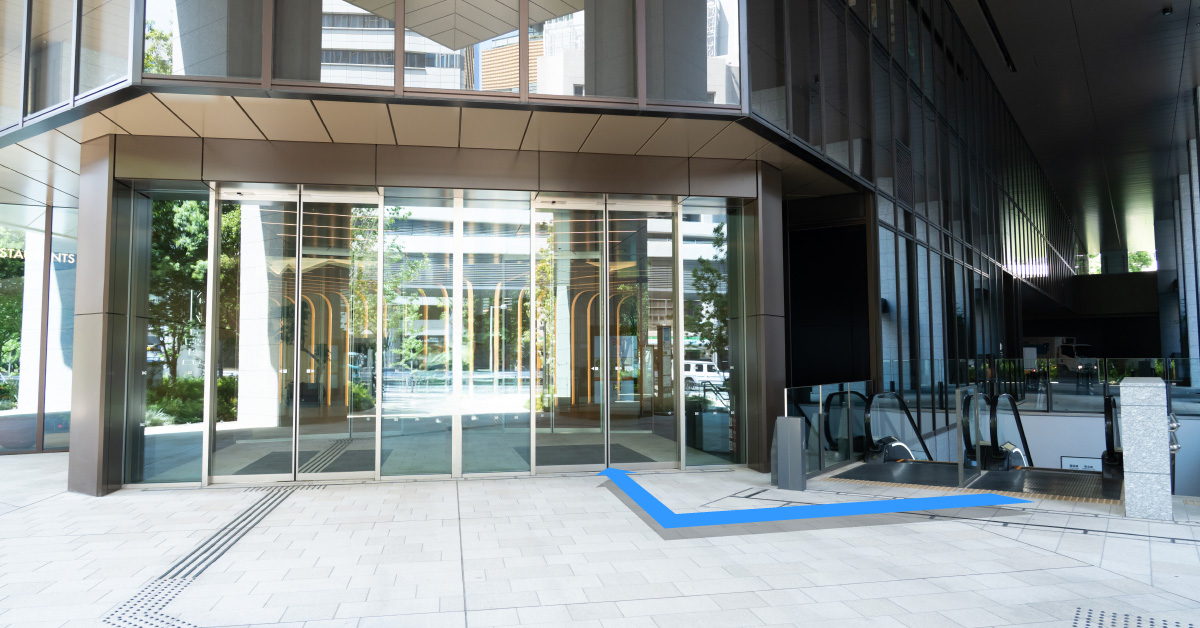 正面にガラス扉の入口、右側にエスカレーター。青い矢印がエスカレーターから入口に向かう経路を示す