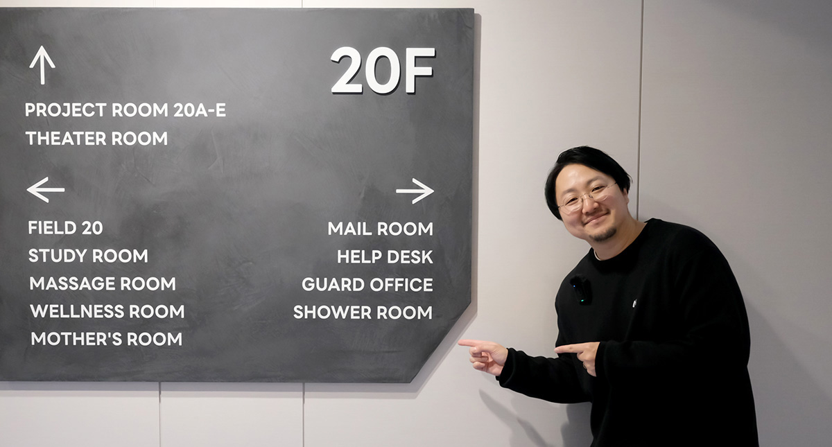 佐藤が白い壁の前に設置されたグレーの案内サインボードの横に立ち、両手でボードを指差しながら笑顔を向けている