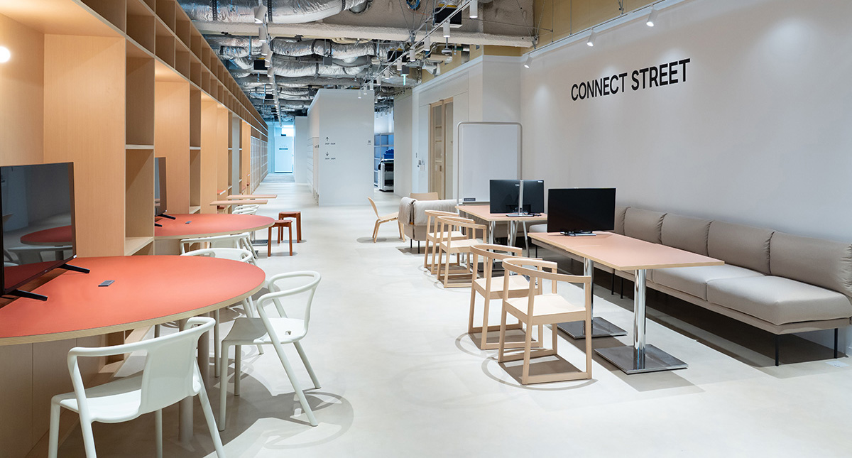オフィス内の共有スペースを奥行き方向に捉えた写真。右側の白い壁には「CONNECT STREET」という黒い文字が大きく表示され、その下にベージュ色のソファと木製テーブル、モニターが設置されている。左側には木目調の棚が連なり、赤い円形テーブルと白い椅子、モニター付きの席が並んでいる