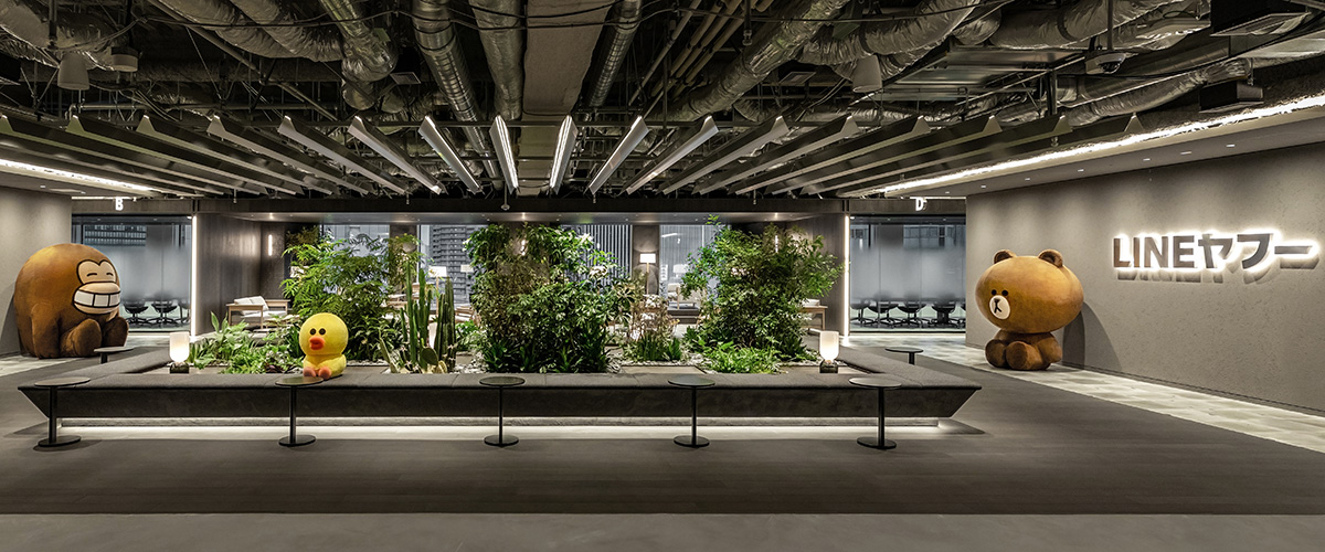 天井にダクトや配管がむき出しになった広いエントランス空間の写真。中央には大きな植栽スペースが設けられ、さまざまな観葉植物やサボテンが立体的に配置されている