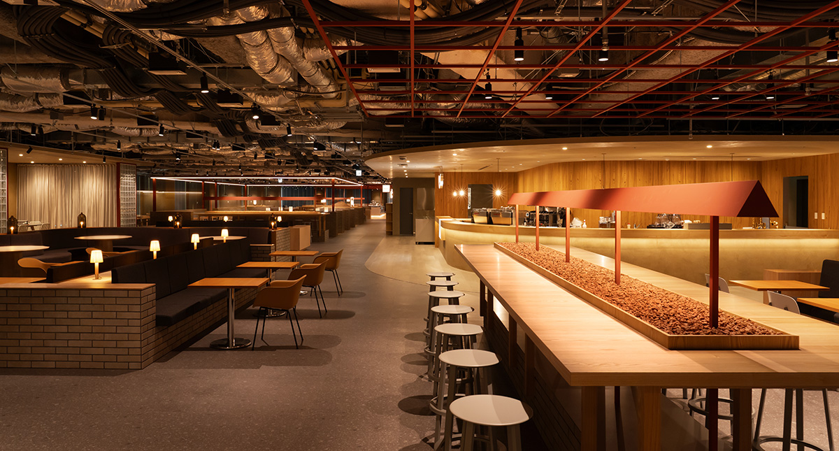 ダイニングエリアを斜め方向から捉えた写真。右側には長い木製のハイテーブルが伸び、その中央に赤いフレーム状の照明と装飾用の小石が敷かれた帯状スペースが設けられている