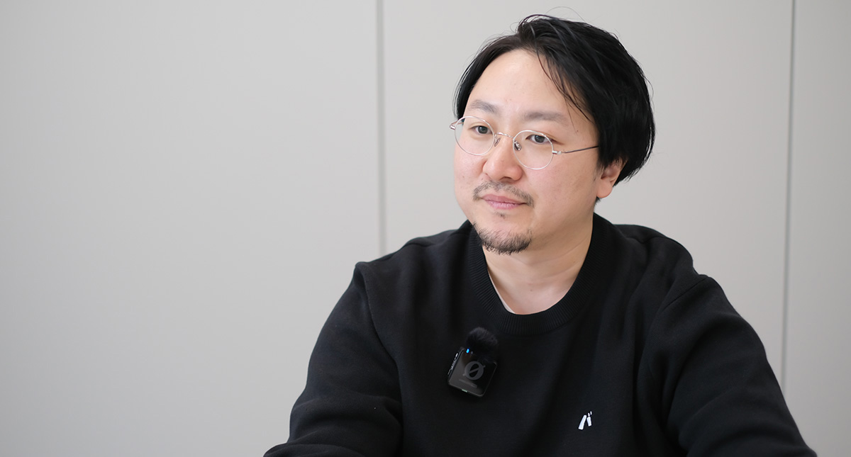 佐藤が机に向かって体をやや横に向け、落ち着いた表情で相手の話を聞いている様子