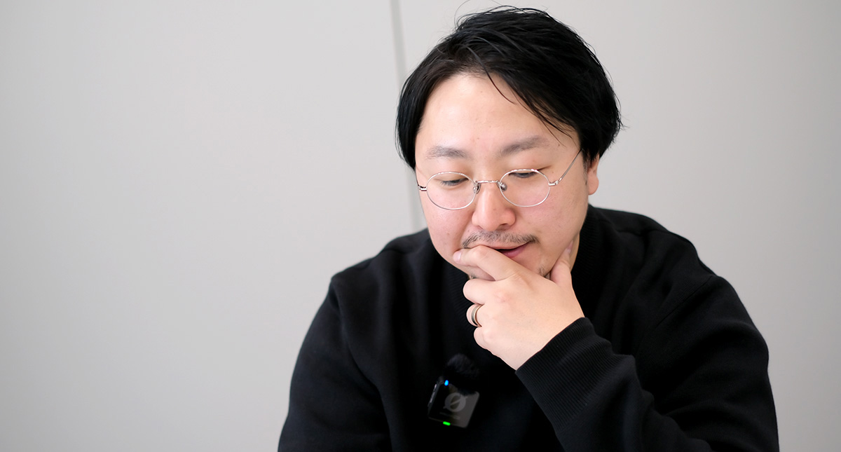 佐藤が机に向かってややうつむき、右手であごに手を当てながら物思いにふけるような表情をしている