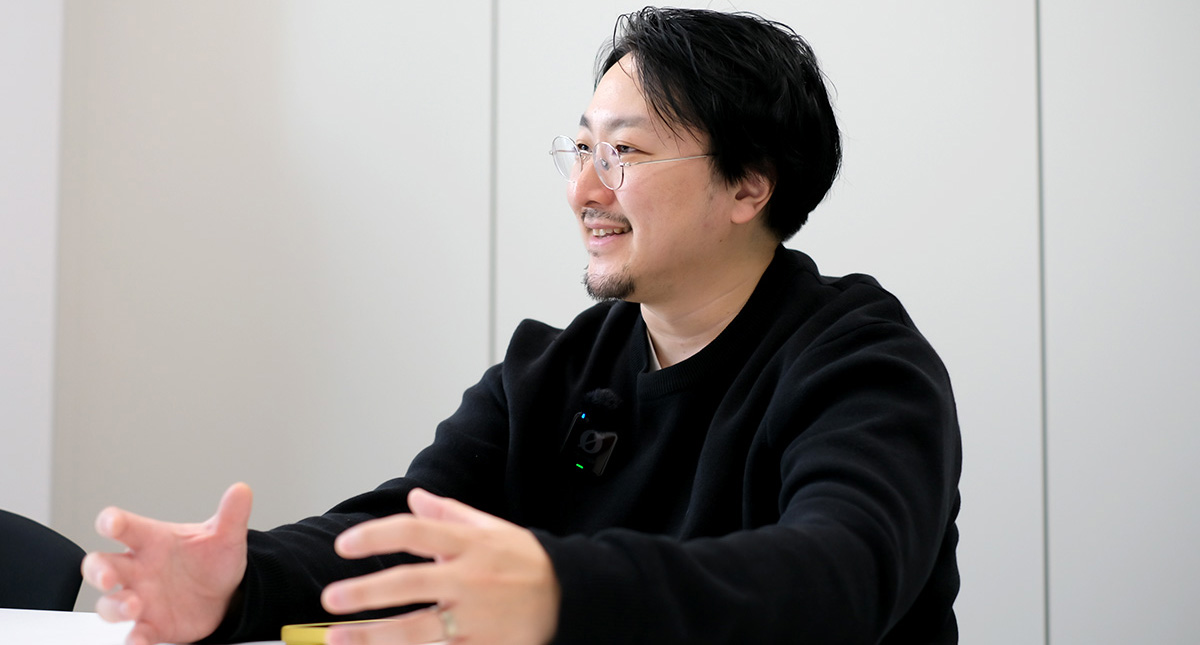 佐藤のバストショットです。机に向かって体をやや横向きにしながら、両手を前に広げて身振りを交えて話している様子