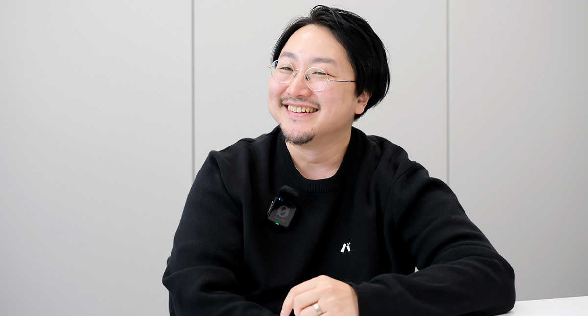 佐藤が机に向かって体をやや横に向け、両腕をテーブルに置きながら明るい笑顔を浮かべている
