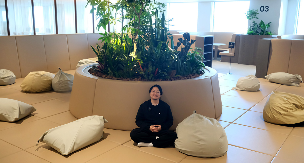 佐藤がベージュ色のクッションフロアの上にあぐらをかいて座り、両手を軽く組んでリラックスした表情を浮かべている。背後には円形の大きな植栽スペースがあり、中央に背の高い観葉植物やサボテンが茂っている