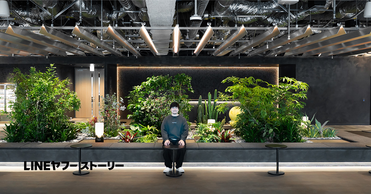 オフィス空間の中央に設けられた大きな植栽スペースの前で、佐藤（男性）が座っている。背景には多種多様な観葉植物やサボテンが配置され、間接照明で縁取られた壁面と、天井にむき出しのダクトや照明器具が並ぶモダンなオフィス内装が広がっている。