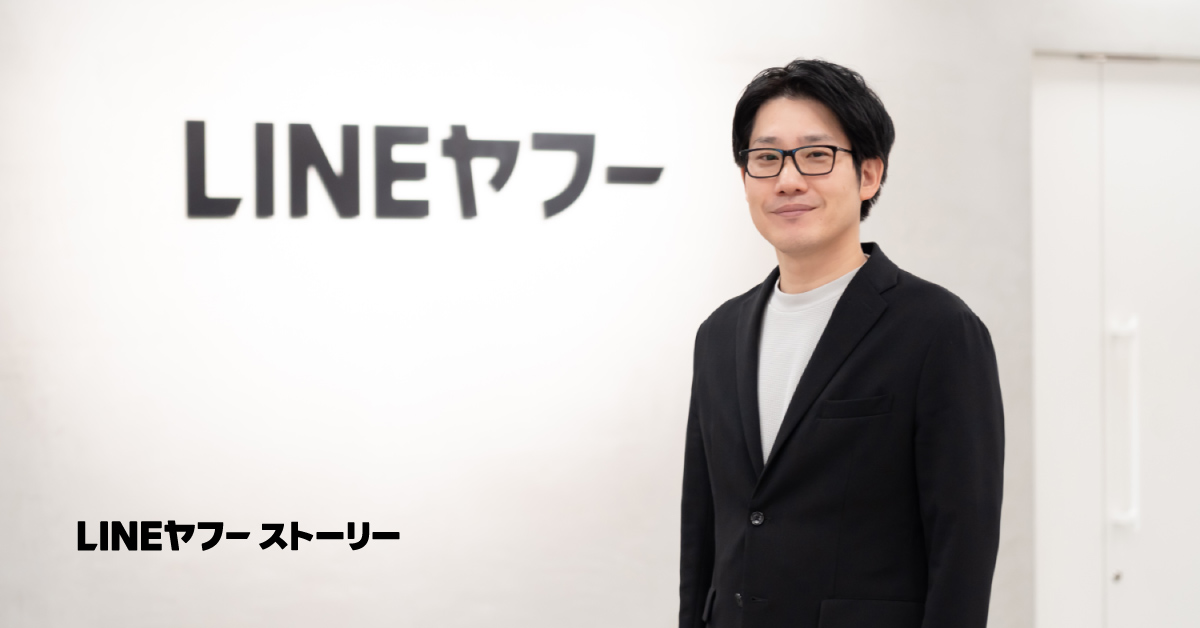 白い壁を背景に「LINEヤフー」というロゴが掲げられているオフィス空間で、林がカメラの方を向いて立っている。黒のジャケットの下に白いインナーを着用し、黒縁の眼鏡をかけている。