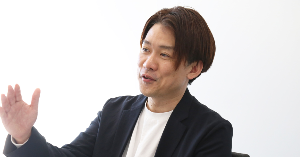 宮本が室内で話しながら左手を上げてジェスチャーしている写真。黒いジャケットに白いTシャツを合わせ、センター分けのやや長めの茶色がかった髪型をしている。