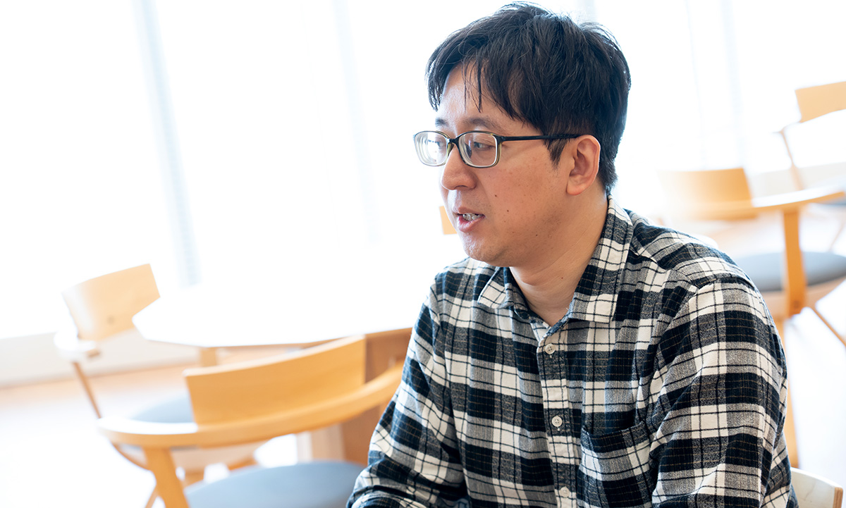 海老原のバストショットです。短い黒髪で眼鏡をかけ、白と黒のチェック柄シャツを着用している。やや横を向きながら口を開き、話している様子。