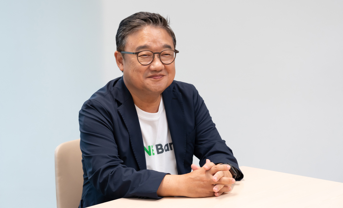 眼鏡をかけたファンが、室内のシンプルな背景の前で机に向かって座っている写真。男性はネイビーのジャケットに白い「LINE Bank」と書かれたTシャツを着用しており、両手を軽く組んで机の上に置いている。短く整えられたグレーがかった髪型で、穏やかな表情を浮かべている。