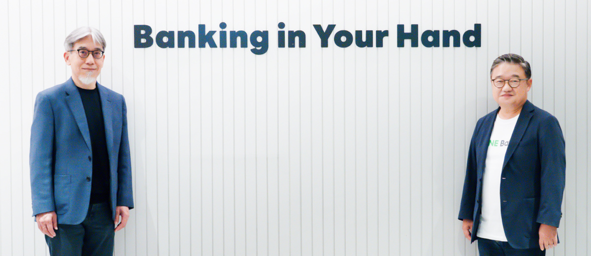 モリスとファンが、白い縦縞の壁を背景に左右に離れて立っている写真。壁の上部中央には「Banking in Your Hand」という文字が大きく掲げられている。