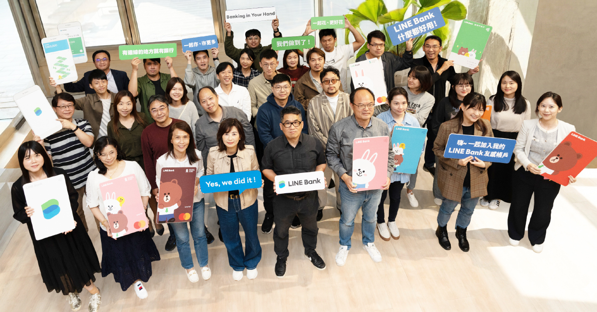 多くの男女が室内に集まり、カメラを見上げるようにして立っている集合写真。参加者はLINE Bankのロゴ入りボードや、キャラクターが描かれたパネル、スローガンが書かれたサインボードを手にしている。「Banking in Your Hand」「Yes, we did it!」「LINE Bank」などの英語のほか、中国語で書かれたメッセージも掲げられている。
